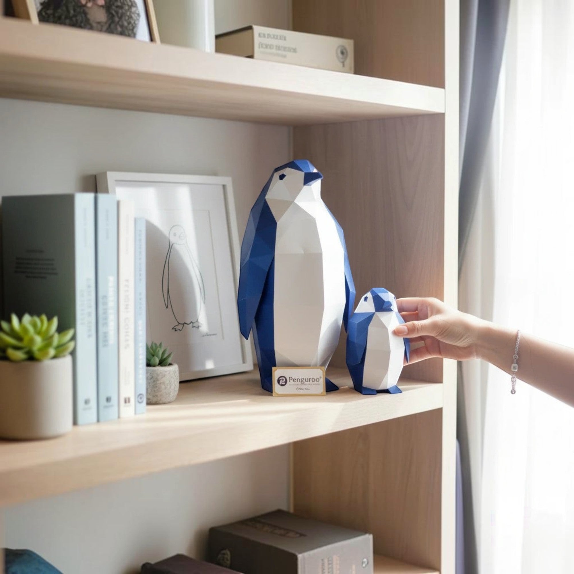 Penguroo® Gifts Inc. set of two geometric penguins, one large and one small, in blue and white, displayed on a shelf with books and plants.